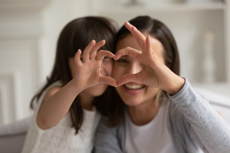Happy young mother with cute little daughter making focused heart sign with hands, looking at camera. Smiling millennial mom and small girl showing love gesture together, expressing care, affection.