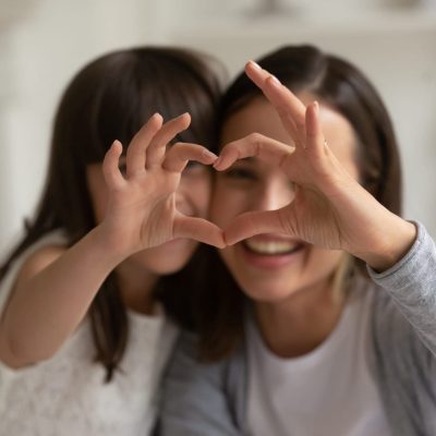 Happy young mother with cute little daughter making focused heart sign with hands, looking at camera. Smiling millennial mom and small girl showing love gesture together, expressing care, affection.