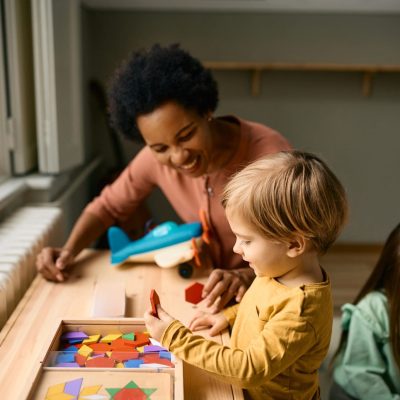 Happy boy playing with colorful puzzle shapes with his kindergarten teacher.