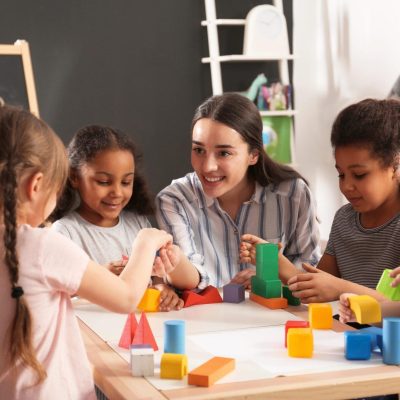 Cute little children and nursery teacher playing with building blocks in kindergarten. Indoor activity