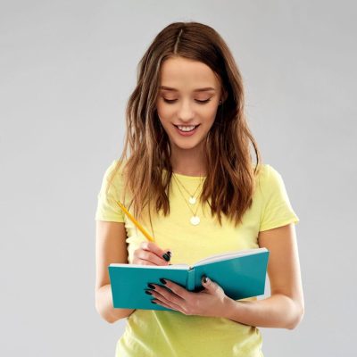 education, school, inspiration and people concept - smiling young woman or teenage student girl in yellow t-shirt writing to diary or notebook by pencil over grey background