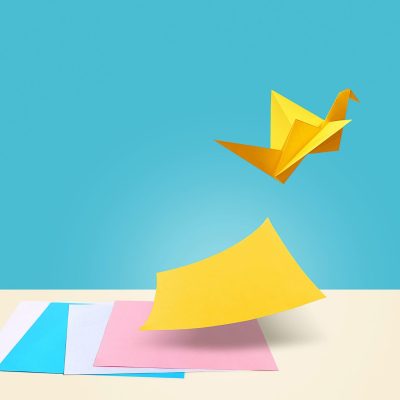 There are four colorful papers on a table with a blue background. Yellow paper is the symbol for creative ideas which is separating itself from other papers by flying like a bird.